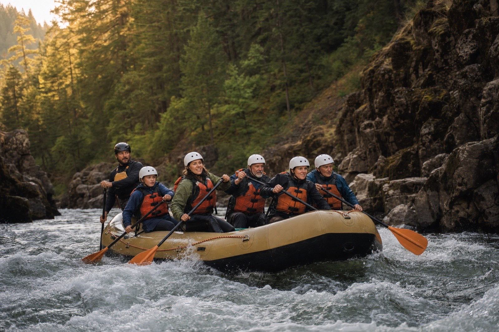 A guide and six guests paddling a raft through moderate whitewater in a forested basalt canyon