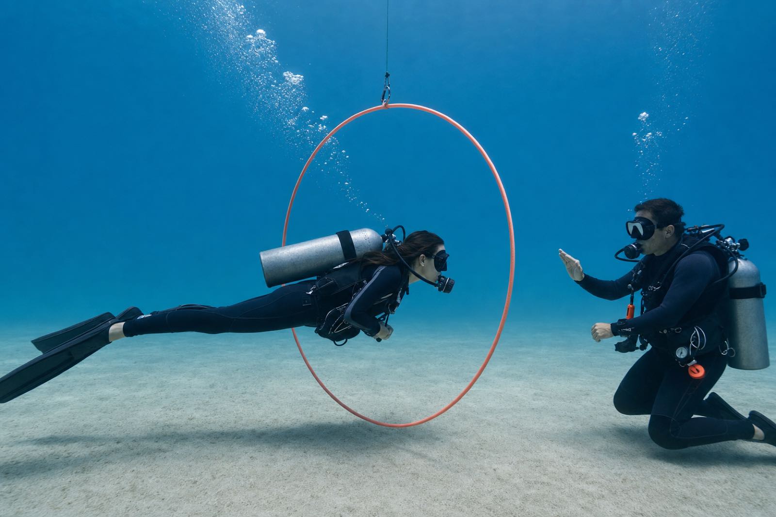 A certified diver practicing neutral buoyancy through a suspended training hoop while an instructor observes