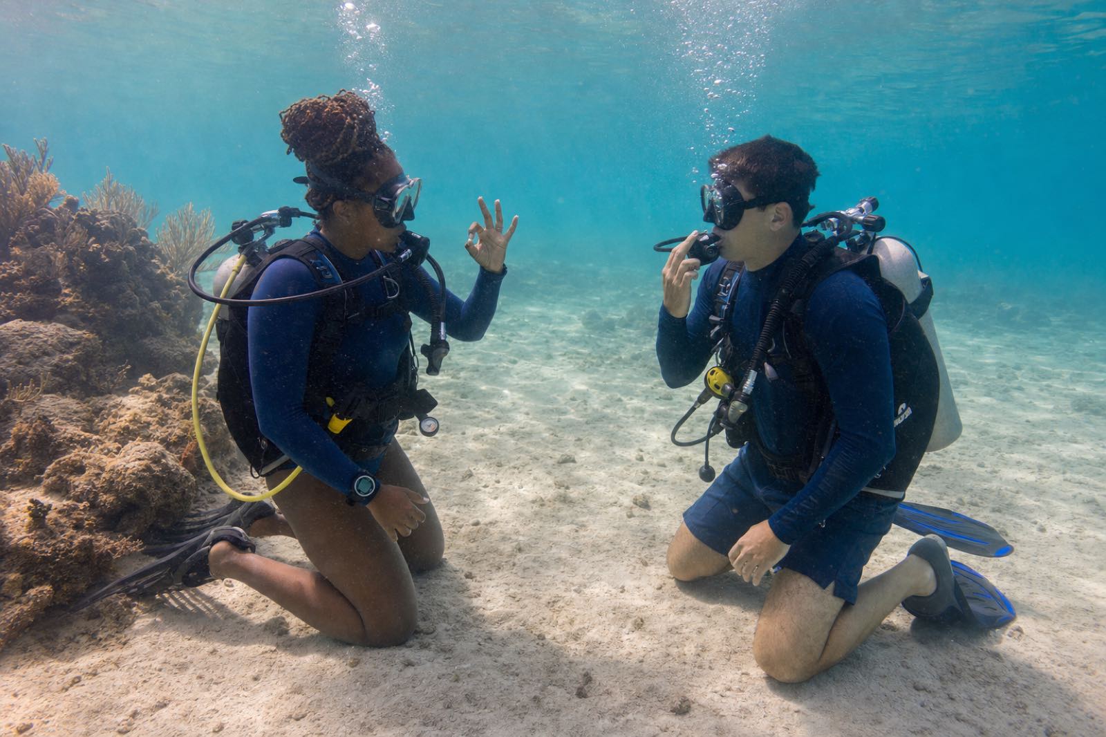 A scuba instructor calmly demonstrating regulator breathing and an OK signal to a first-time diver in shallow water