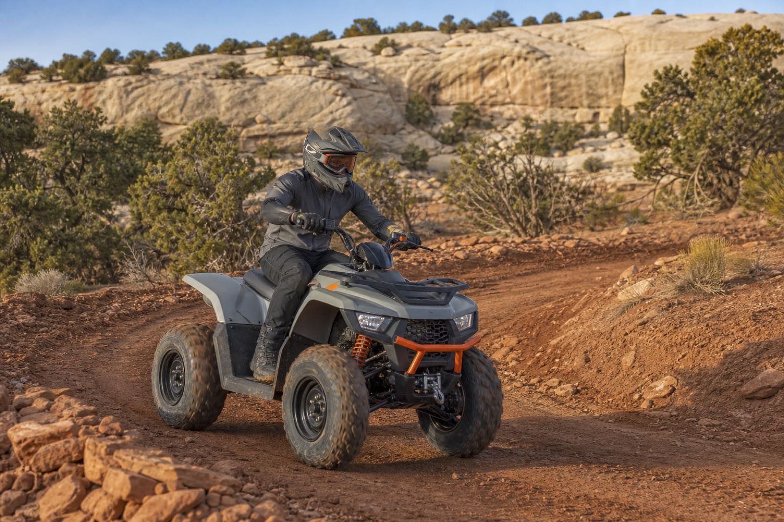 A fully equipped rider guiding a midsize ATV through a controlled red-clay switchback below sandstone shelves