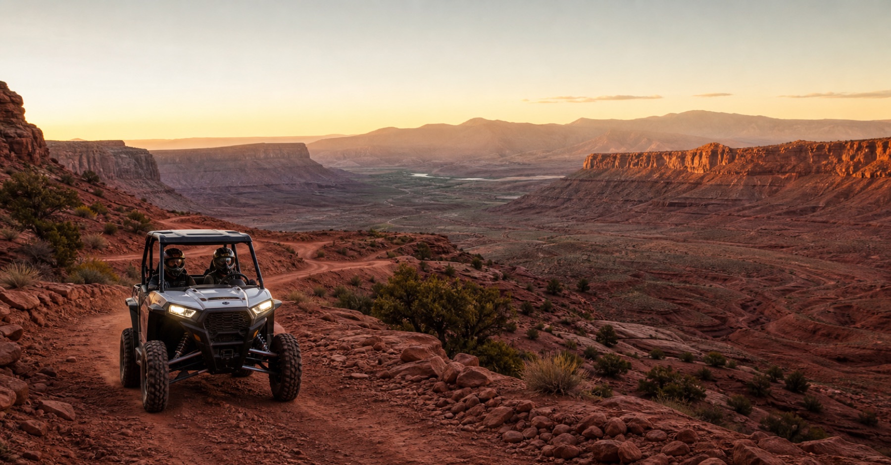 Two helmeted riders in a UTV on a red rock switchback at sunset